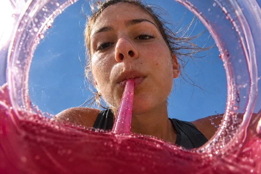 Mulher bebendo com canudo rosa de um copo transparente cheio de líquido rosado, vista de baixo contra o céu azul